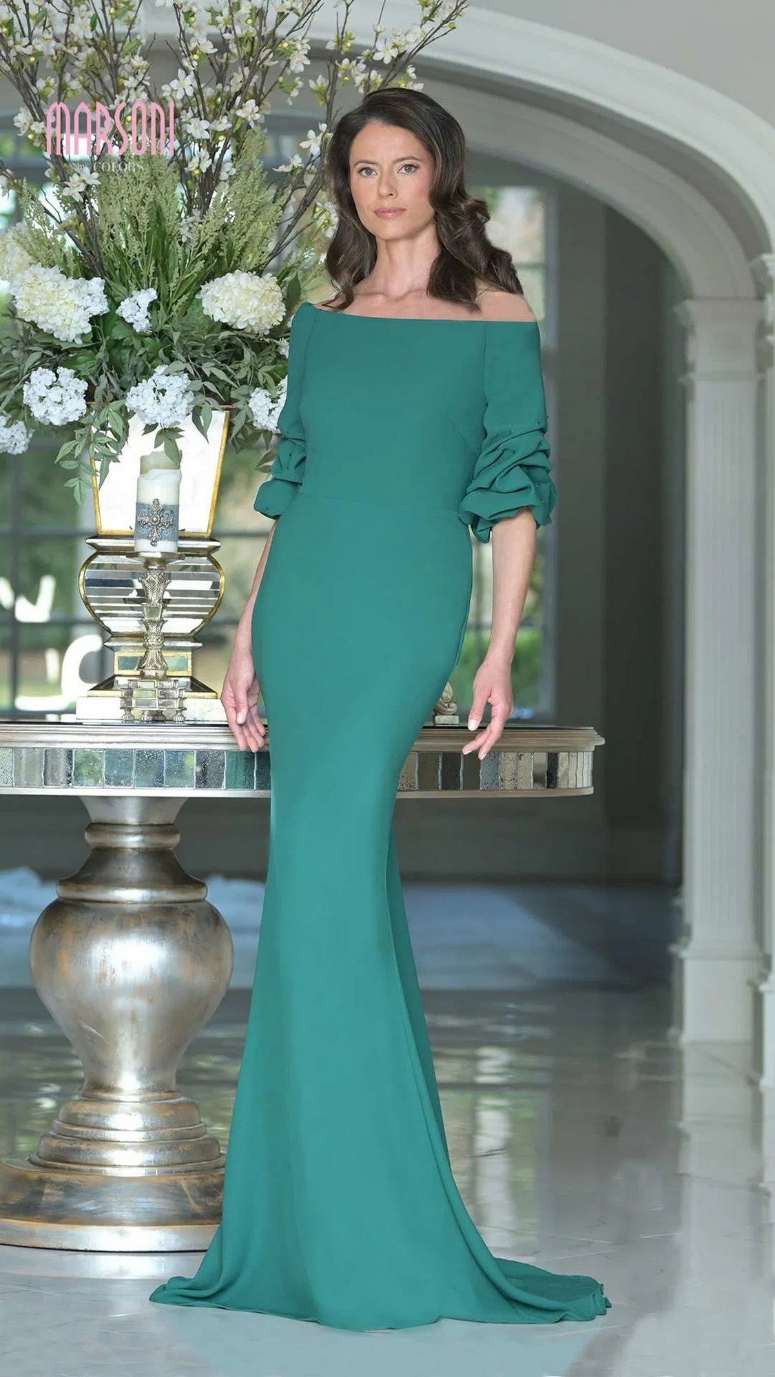 A woman stands indoors in a fitted, emerald Marsoni MV1337 long formal dress with ruched off-the-shoulder sleeves, posed beside a table topped with a tall floral arrangement.